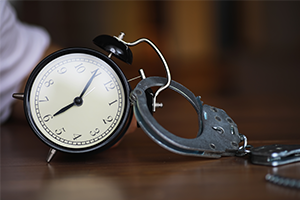 An alarm clock attached to a pair of metal handcuffs rests on a wooden surface, creating a playful yet unsettling image about time and confinement.