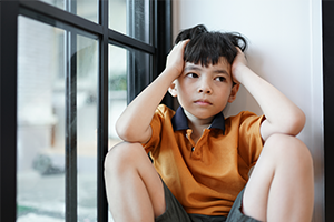 A young boy in an orange shirt sits by a large window, holding his head and looking worried or overwhelmed in the quiet room.