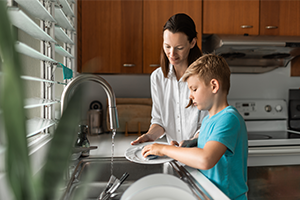 A mother and young son wash dishes together at a kitchen sink, sharing a calm, warm moment in a sunlit, wood-cabinet kitchen.
