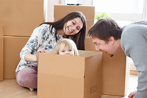 A smiling family peeks into a cardboard moving box: a mother and toddler inside, with the father leaning close, creating a playful, happy moving-day scene.
