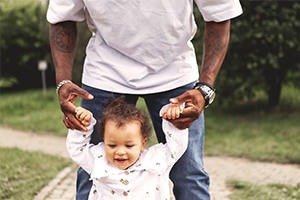 A smiling toddler in a white outfit takes steps outdoors while an adult gently holds both hands; a grassy park setting conveys a joyful, supportive moment.