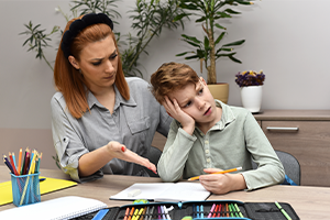 A concerned teacher gestures toward an open workbook as a tired-looking student rests his cheek on his hand, pencil in hand, at a classroom desk.