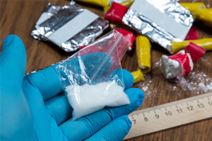 A blue-gloved hand holds a small bag of white powder beside foil packets and a ruler on a wooden table, suggesting forensic evidence collection.